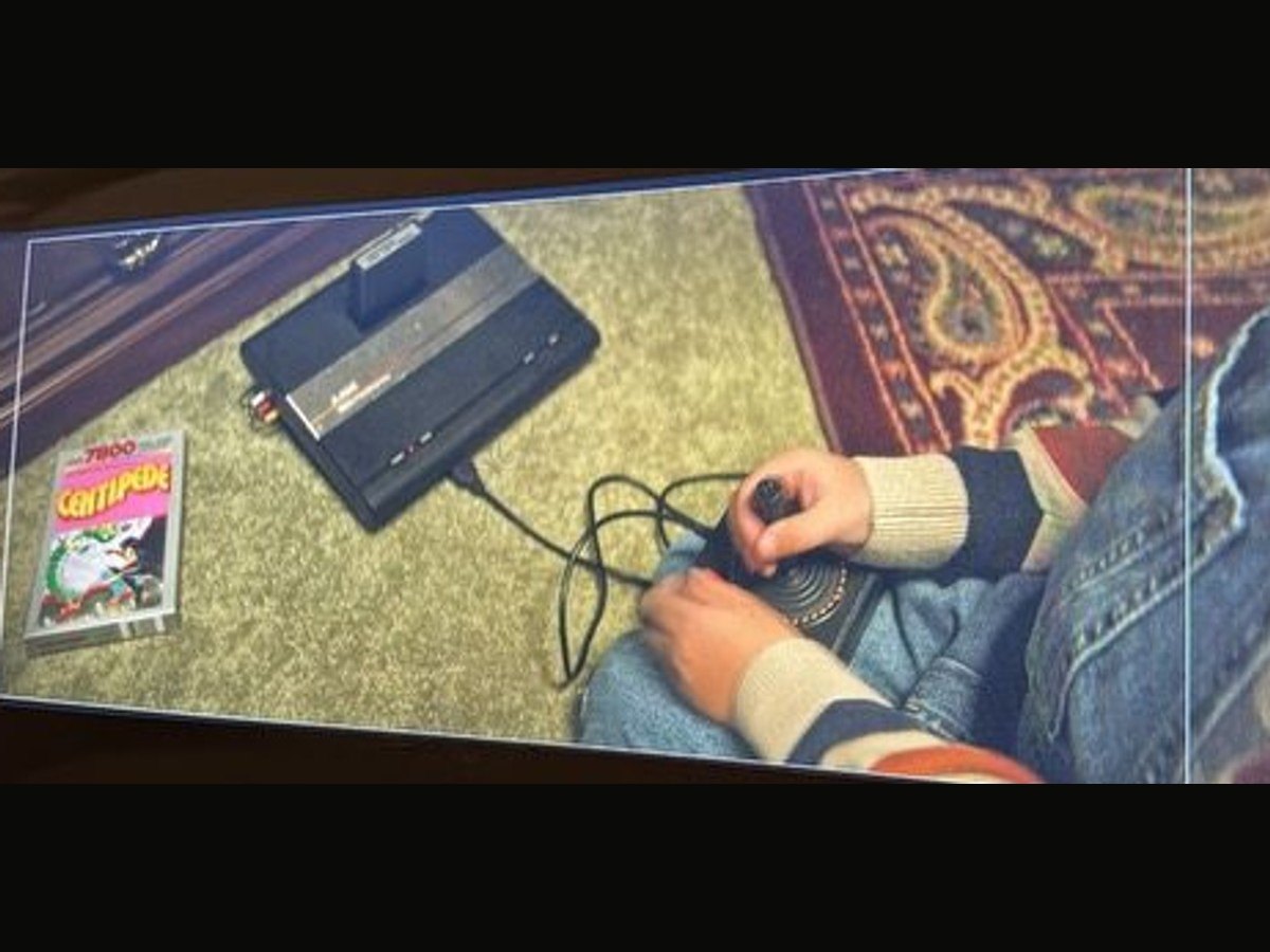 An Atari 7800 console and a boxed Centipede cartridge on a green carpet, a child's hands on the joystick